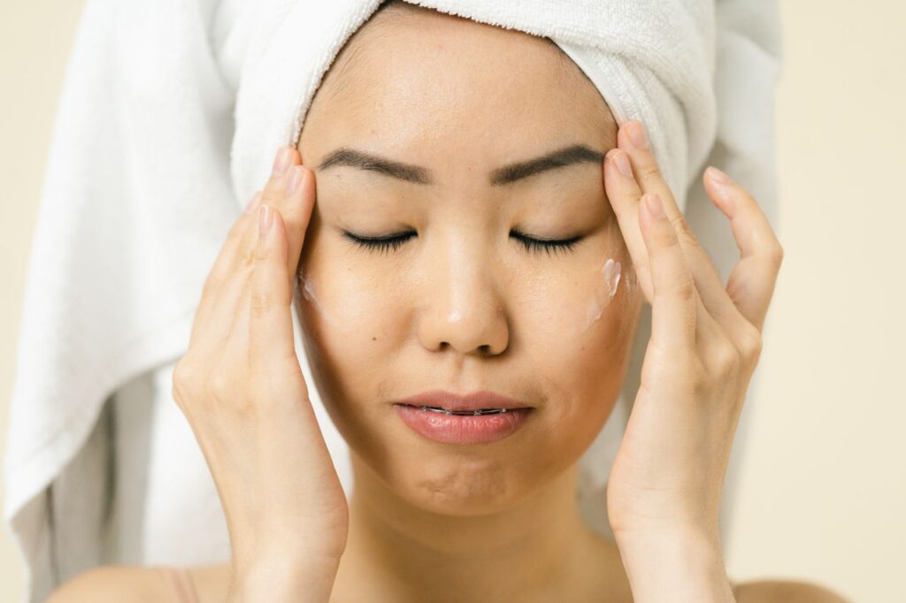 Asian woman with a towel on head applying moisturizer for skincare routine.